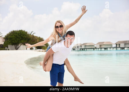 Ritratto di uomo sorridente donna portante sulla sua schiena lungo la riva del mare. Guy dando piggyback ride alla ragazza dall' oceano. Coppia giovane avendo divertimento su Foto Stock