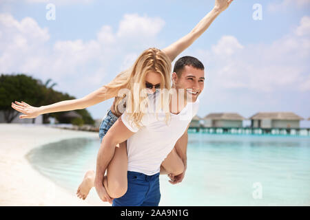 Ritratto di uomo sorridente donna portante sulla sua schiena lungo la riva del mare. Guy dando piggyback ride alla ragazza dall' oceano. Coppia giovane avendo divertimento su Foto Stock