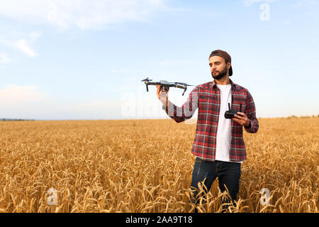 Tanga barbuto uomo mostra un piccolo drone compatto e mantiene il dispositivo di controllo remoto nella sua mano. Imprenditore agronomo guarda quadcopter prima del lancio in un Foto Stock