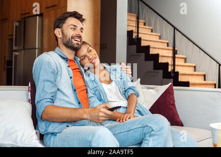Razza mista matrimonio. Giovane uomo e donna abbracciando la visione di commedia in tv insieme a ridere allegro Foto Stock