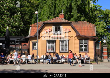 Una donna e bambino a piedi passato persone sedute al sole durante la stagione estiva, fuori Grindstugan café e bar a Gothenburg, Svezia. Foto Stock