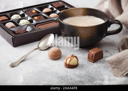 Casella di lusso di cioccolatini selezione con tazza di cappuccino e caffè cucchiaio d'argento su sfondo chiaro. Foto Stock