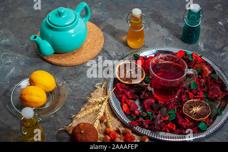 Red Hot Hibiscus tè in una tazza di vetro su di un tavolo di legno tra i petali di rosa e tè secco crema pasticcera con carcade Foto Stock