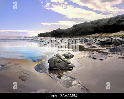 Dettaglio di una lava spiaggia di sabbia con drenaggio di acqua all'Oceano Atlantico su Tenerife. In primo piano una grande pietra lavica con focus sulla pietra. Blue Foto Stock