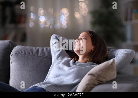 Donna felice rilassante con le braccia sulla testa seduto su un divano nella notte a casa Foto Stock