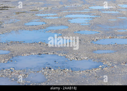 Sole sul parcheggio hardcore con pozze d'acqua in piedi in cavità / pozze tuffi. Cielo blu riflesso in pozze, riflessi acqua. Foto Stock