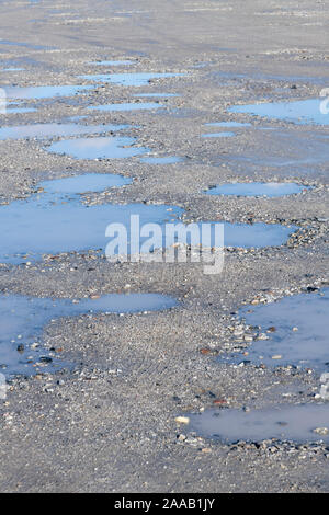 Sole sul parcheggio hardcore con pozze d'acqua in piedi in cavità / pozze tuffi. Cielo blu riflesso in pozze, riflessi acqua. Foto Stock