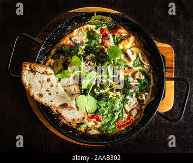 Frittata spagnola prima colazione servita calda in padella con spinaci, funghi, pomodori, formaggi, pane Foto Stock