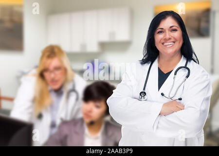 Bello ispanico medico o infermiere in piedi nel suo ufficio con il personale che lavora dietro. Foto Stock