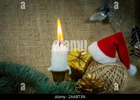 Sfondo di natale con candela che brucia, decorazioni, dono, rotolo di spago e Santa hat su tela rustico sfondo con copia spazio per una vacanza gre Foto Stock