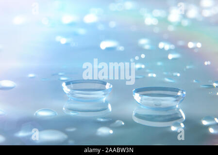 Lenti a contatto su vetro piano riflettente con gocce di acqua e isolato sfondo blu. Composizione orizzontale. Vista in elevazione frontale. Foto Stock