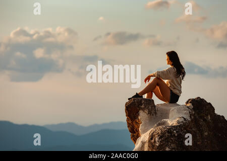 Una ragazza sportiva si siede sul big rock e guarda al Cielo di tramonto e montagne Foto Stock