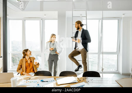 Il gruppo di un giovane creative office dipendenti aventi una certa discussione informale, mentre in piedi insieme nella luminosa sala meeting con grandi finestre Foto Stock