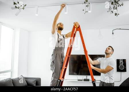 Due repairmen o professional elettricisti installazione di punti luce, stando in piedi sulla scala nel salotto del moderno appartamento Foto Stock