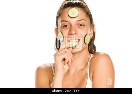 Giovane donna sorridente in posa con fette di cetrioli sul suo viso su sfondo bianco Foto Stock