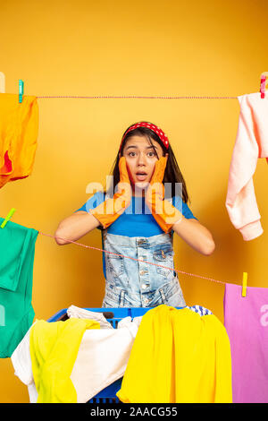 Divertente e bella casalinga a fare i lavori di casa isolata su sfondo giallo. Giovane donna caucasica circondato da lavare i vestiti. La vita domestica, grafica brillante e le pulizie del concetto. Scioccato. Foto Stock