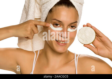 Giovane donna sorridente l'applicazione di crema per il viso sotto i suoi occhi su sfondo bianco Foto Stock