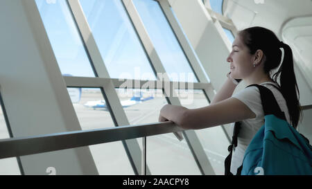 Donna felice turista sta parlando al telefono cellulare nel terminale di un aeroporto in area di attesa. Ella è in possesso di documenti sulle sue mani e guardando alla finestra, vista laterale. Foto Stock