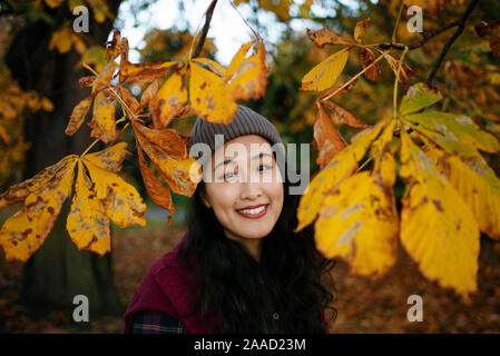 RF Close-up testa di sorridente asiatico (giapponese) femmina nel parco tra foglie di albero. Tema autunnale, RM disponibile. Novembre 2019 Foto Stock