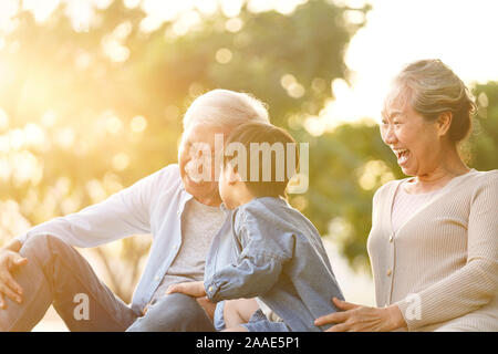 Nipote di asiatici, il nonno e la nonna seduta su erba avendo divertimento all'aperto nel parco al tramonto Foto Stock