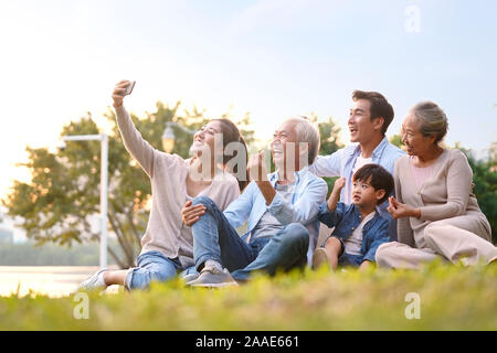 Tre generazioni felice famiglia asiatica seduto sull'erba prendendo un selfie utilizzando il telefono cellulare all'aperto nel parco Foto Stock