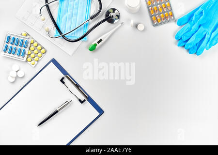 Attrezzatura medica : pillole, maschera, guanti blu, termometro e uno stetoscopio, bianca vuota con una penna su sfondo bianco. Vista dall'alto. Spazio di copia Foto Stock