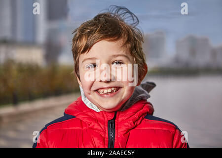 Carino sorridente 7 anni bambino in vestiti caldi su una strada di città nel periodo invernale. La faccia di un allegro ragazzo che cammina in un rosso down jacket in weat freddo Foto Stock