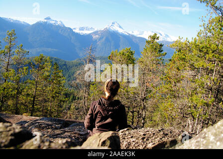 Un naturale giovane donna con marrone capelli intrecciati, seduto ancora sulle rocce in una postura contemplativa, fissando il Snow capped tantalo mountain range Foto Stock