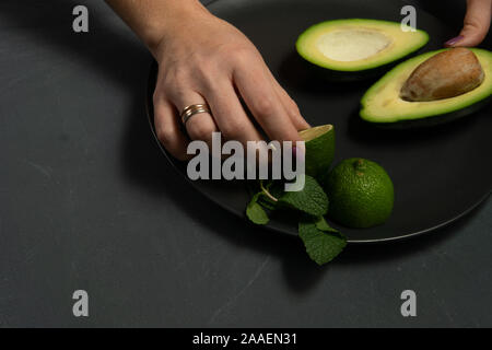 La donna la preparazione di materie di avocado e calce su una piastra nera su sfondi scuri Foto Stock