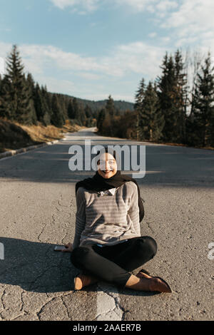 Donna con hijab seduti lungo la linea di segnaletica orizzontale.ella si trova nel centro di una tranquilla strada in montagna,circondato da alberi. Ella è prendere il sole Foto Stock