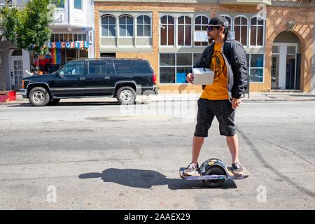 Un uomo detiene una scatola e cavalca un monociclo motorizzato in strada nel quartiere di missione quartiere di San Francisco, California, 18 luglio, 2019. () Foto Stock