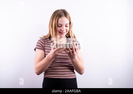 Caucasian ragazza adolescente che indossa un colorato a strisce orizzontali di T-shirt e sorridere mentre guardando uno smartphone contro uno sfondo bianco Foto Stock