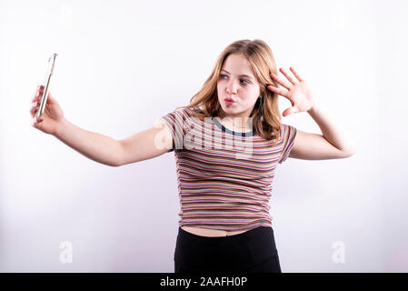 Caucasian ragazza adolescente che indossa un rigato orizzontale T-shirt prendendo un drammatico selfie con uno smartphone contro uno sfondo bianco Foto Stock