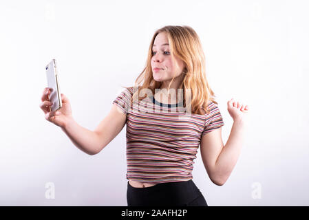 Caucasian ragazza adolescente che indossa un colorato a strisce orizzontali T-shirt con un espressione divertente e pursed lips tenendo un selfie con uno smartphone Foto Stock