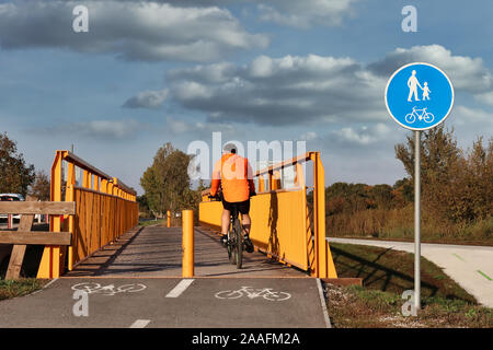 Giovane uomo che indossa colpisce giacca arancione godendo di giro in bicicletta su inizio autunno giornata soleggiata, attraversando in acciaio ciclista arancione / ponte pedonale Foto Stock