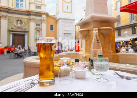 Un cesto di pane e panini, un bicchiere di birra e una bottiglia di acqua su un patio con tavolo in una piazza di fronte a una fontana a Nizza Francia. Foto Stock