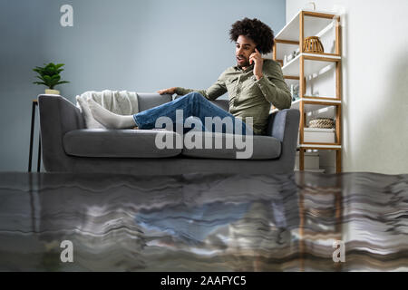 Allagato il pavimento dalla perdita di acqua nella parte anteriore del uomo seduto sul divano chiamando per idraulico Foto Stock