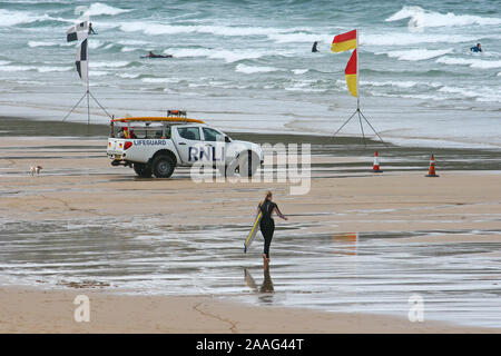 Fistral Bay, Newquay, Inghilterra e una giovane donna capi attraverso la sabbia bagnata per l'acqua con la sua tavola da surf sotto l'occhio vigile dei bagnini. Foto Stock