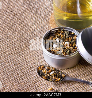 Biologici e naturali riso marrone tè, genmaicha in un contenitore. Accompagnata da una tazza di tè caldo su sfondo marrone. Foto Stock