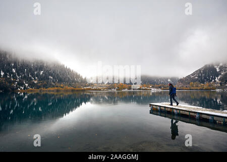 Lonely Man con zaino e fotocamera percorrendo a piedi il molo presso il lago di nebbia con la riflessione in autunno in Kazakistan e in Asia centrale Foto Stock