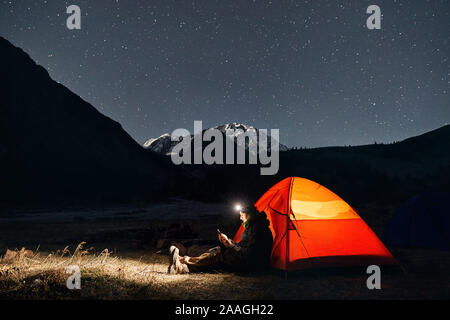 Uomo con proiettore vicino incandescente tenda arancione in montagna sotto il cielo notturno con stelle Foto Stock