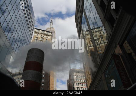 Nuvole di vapore della metropolitana che si innalzano tra i grattacieli di Manhattan, verso l'Empire state Building contro il cielo blu con le nuvole bianche Foto Stock