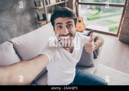 Primo piano della razza mista arab ragazzo seduto comodo divano rendendo selfies sollevando il pollice fino esprimere accordo eccitato indossare abbigliamento casual appartamento loft Foto Stock