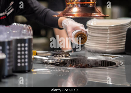 Mani in guanti in lattice tenere la carne fritta con pinze in una padella. Un cuoco prepara la carne in cucina in un ristorante. La cena è quasi pronta. Foto Stock