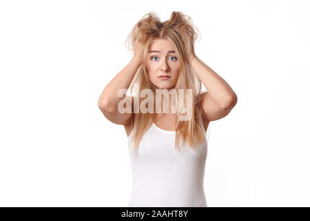 Giovane donna in maglia bianca deluso con il suo confuso e disheveled capelli biondi, in piedi con le braccia alla sua testa, guardando la fotocamera con triste emoti Foto Stock