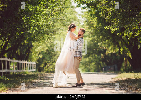 Felice sposo solleva sposa in estate park. uomo sollevato e abbraccio donna per le sue braccia. Appena sposata giovane nel parco. Appena sposato. Passeggiata nel parco Foto Stock