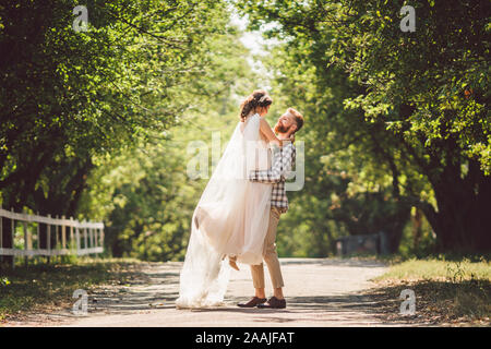 Felice sposo solleva sposa in estate park. uomo sollevato e abbraccio donna per le sue braccia. Appena sposata giovane nel parco. Appena sposato. Passeggiata nel parco Foto Stock