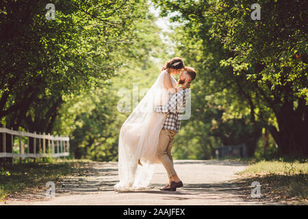 Felice sposo solleva sposa in estate park. uomo sollevato e abbraccio donna per le sue braccia. Appena sposata giovane nel parco. Appena sposato. Passeggiata nel parco Foto Stock