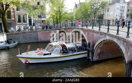 Una barca turistica emerge da sotto un ponte, sul canale Prinsengracht Amsterdam, vicino al suo incrocio con il canale Reguliersgracht Foto Stock