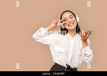 Freestyle. Giovane donna in camicia che indossano le cuffie dancing isolati su bage con lo smartphone per ascoltare musica che mostra gesto di saluto gioioso Foto Stock